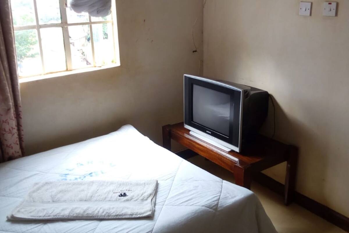 Un dormitorio con cama, televisor sobre un soporte de madera, mosquitero en la ventana y toma de corriente en la pared.
