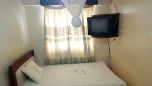 A single bed with a white bedspread and a wooden headboard, a small table, a television mounted on the wall, and a window with sheer curtains. - Salkan Hotel (Embu)