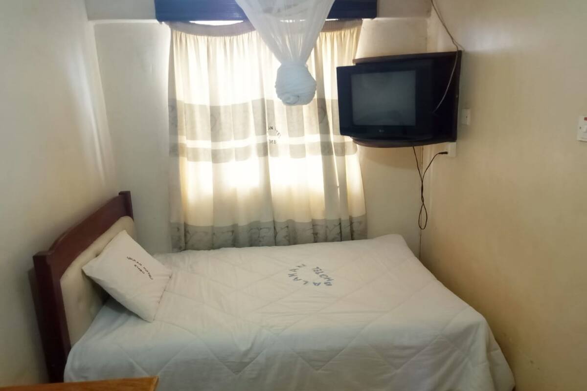Una cama individual con edredón blanco y cabecera de madera, una mesita, un televisor montado en la pared y una ventana con cortinas traslúcidas.