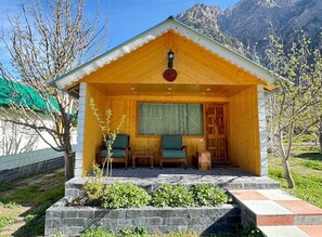 A wooden cabin with a covered porch, two chairs, a small table, and a window. - Banjara Camp and Retreat Sangla (Sangla)