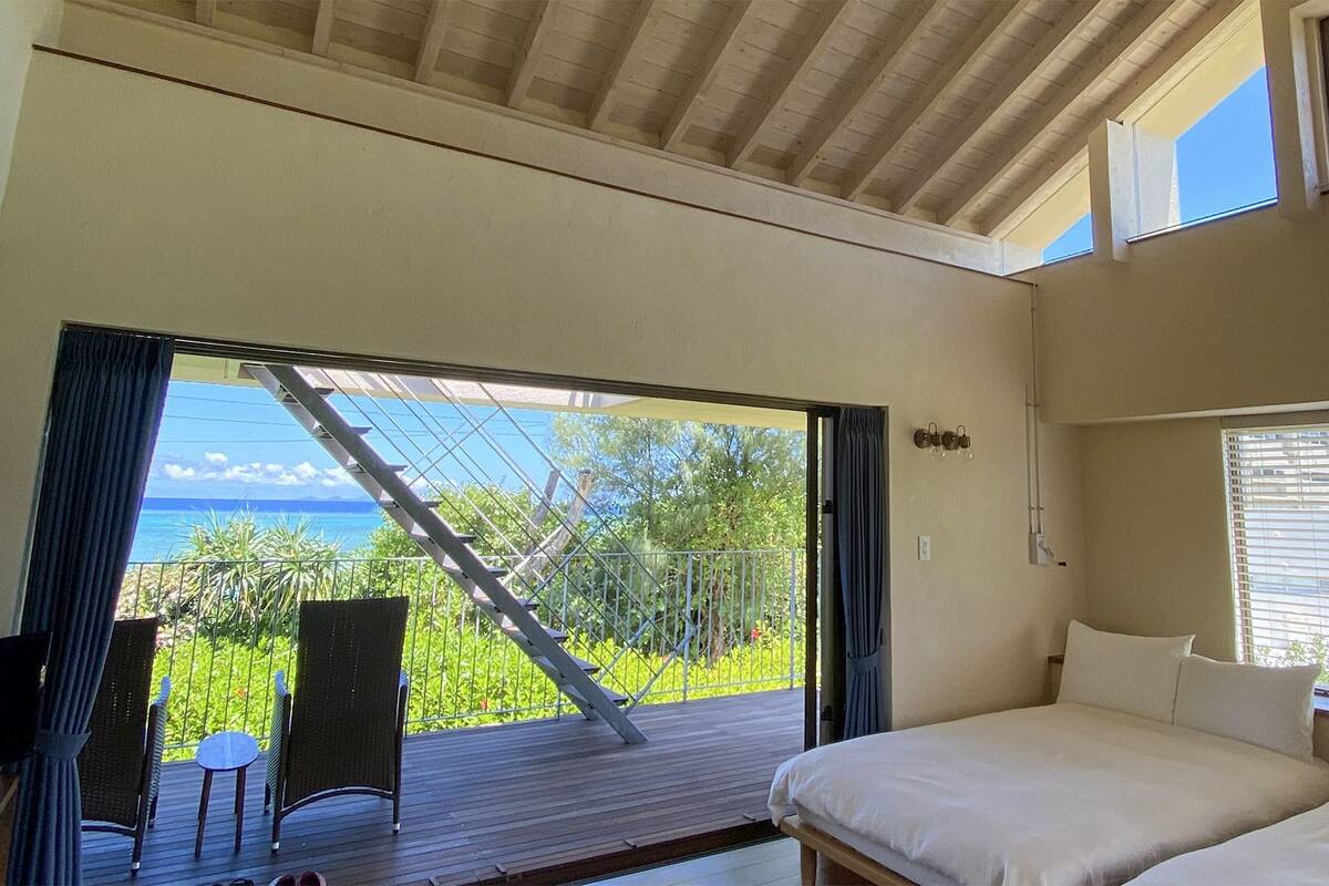 Een slaapkamer met een bed, een stoel, een klein tafeltje en uitzicht op de oceaan door een glazen deur.