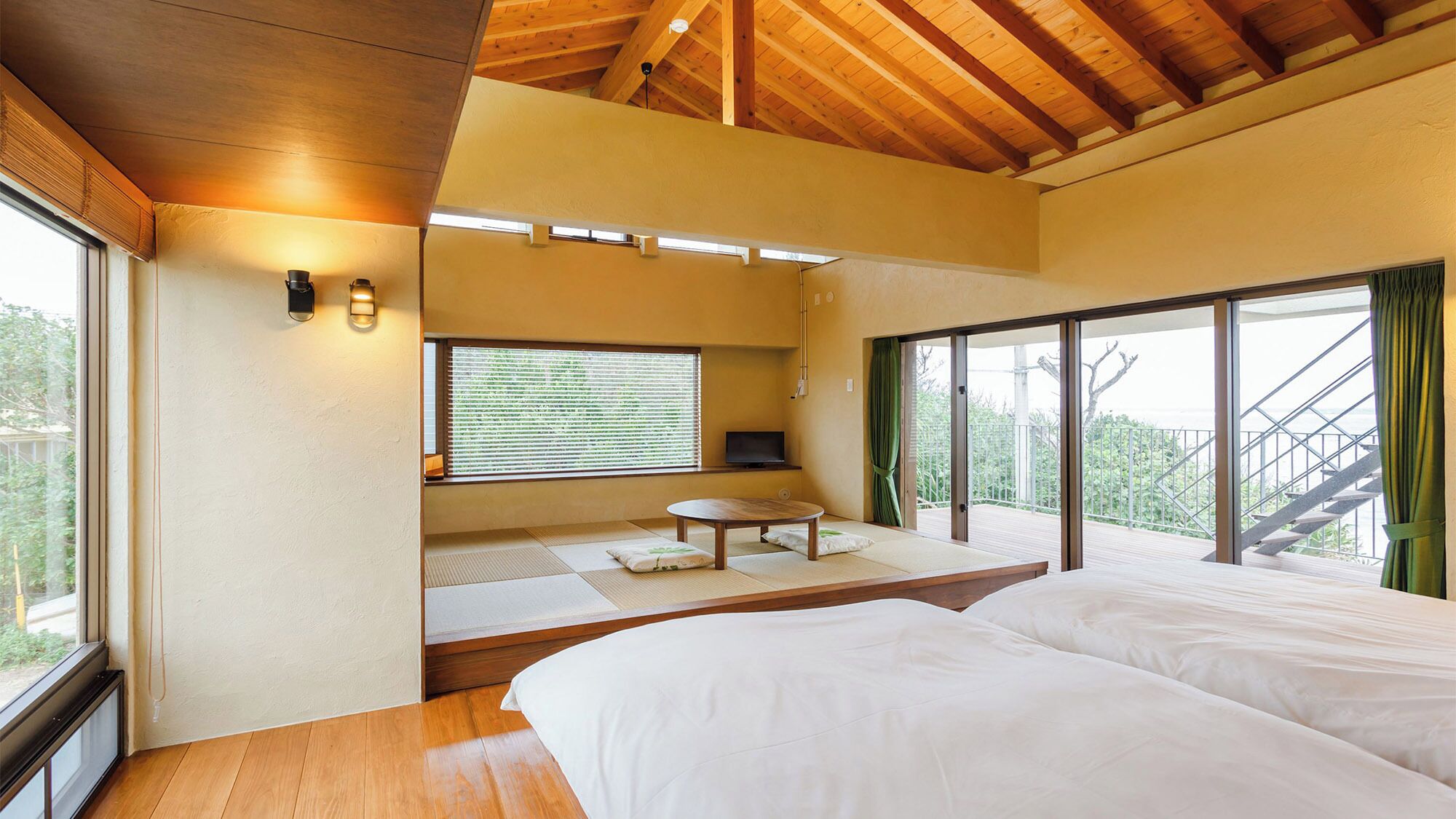 A bedroom with a wooden ceiling, a large window with blinds, a bed, and a small table with cushions.
