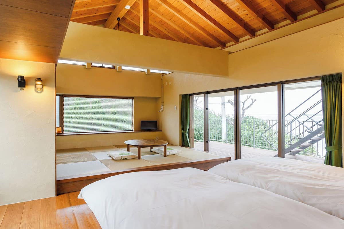 A bedroom with a wooden ceiling, a large window with blinds, a bed, and a small table with cushions.