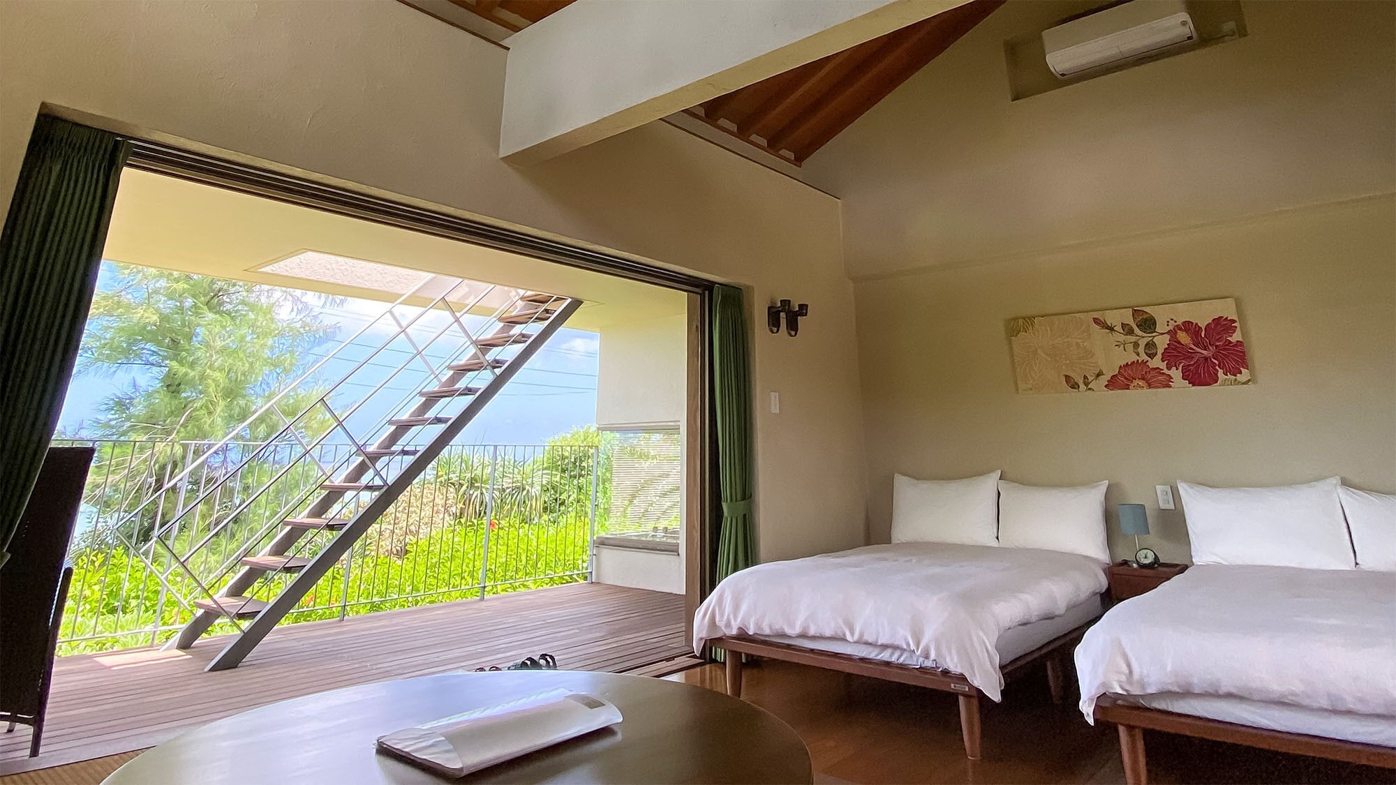 A bedroom with two beds, a wooden table, a TV, and a balcony with a view of greenery and a staircase.