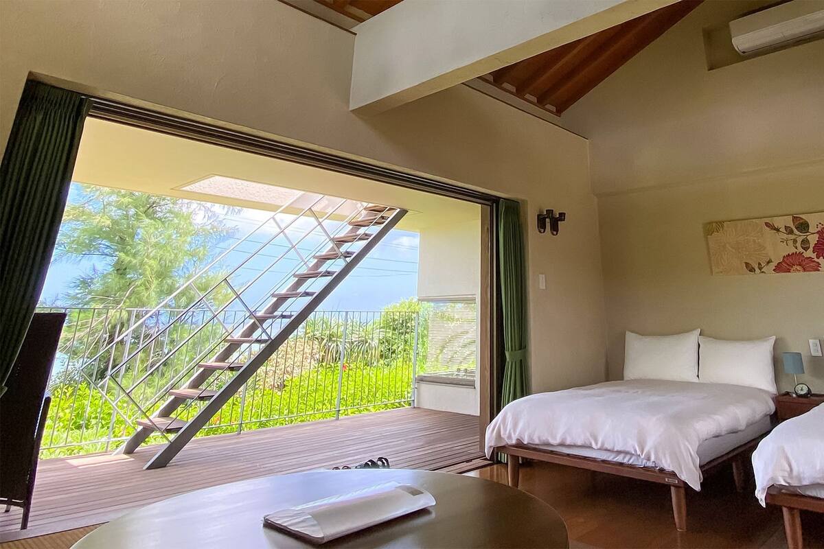 Slaapkamer met twee bedden, een houten tafel, een televisie en een balkon met uitzicht op groen en een trap.