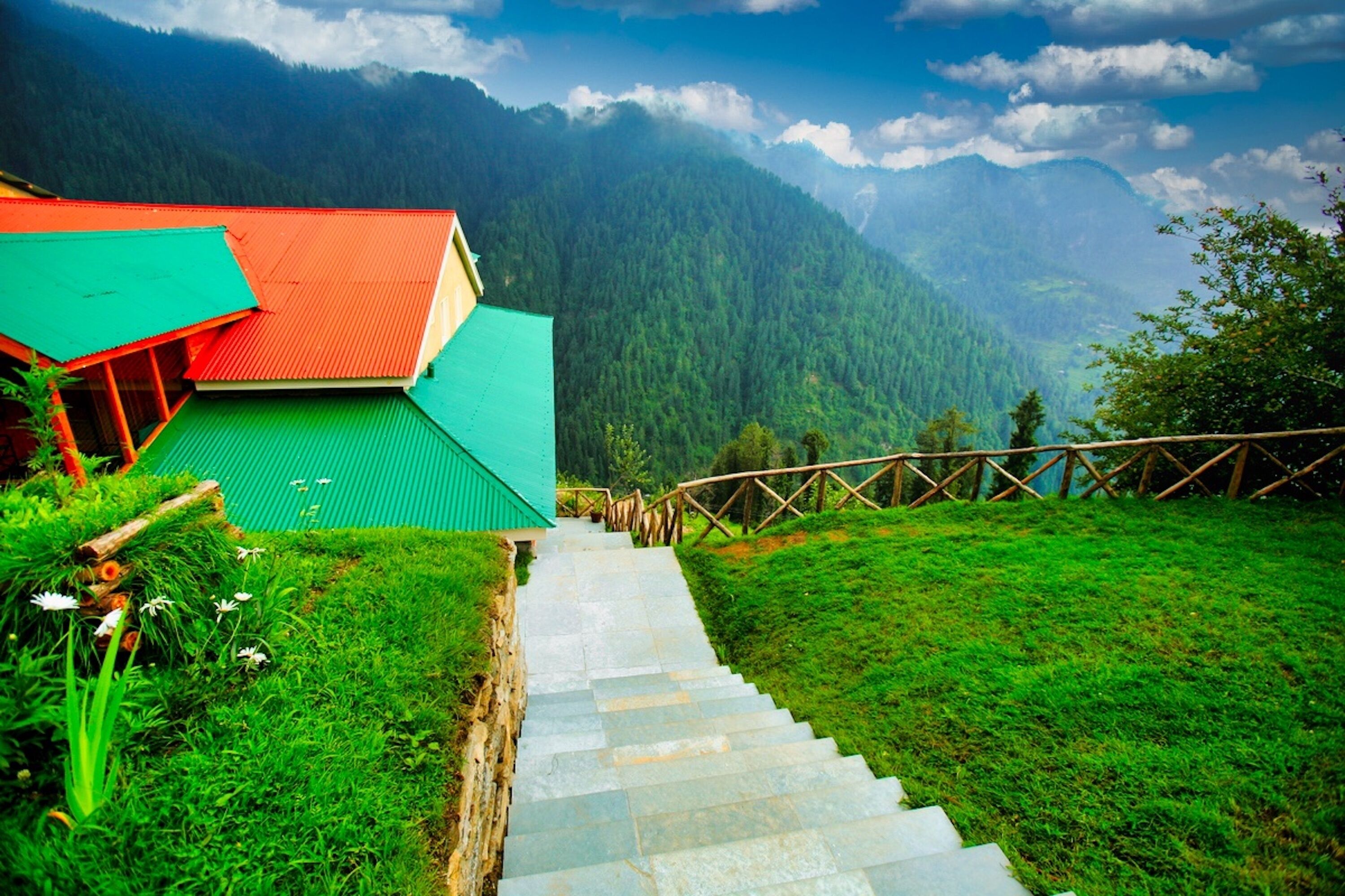A staircase leading to a building with a green roof, surrounded by lush greenery and mountains in the background.