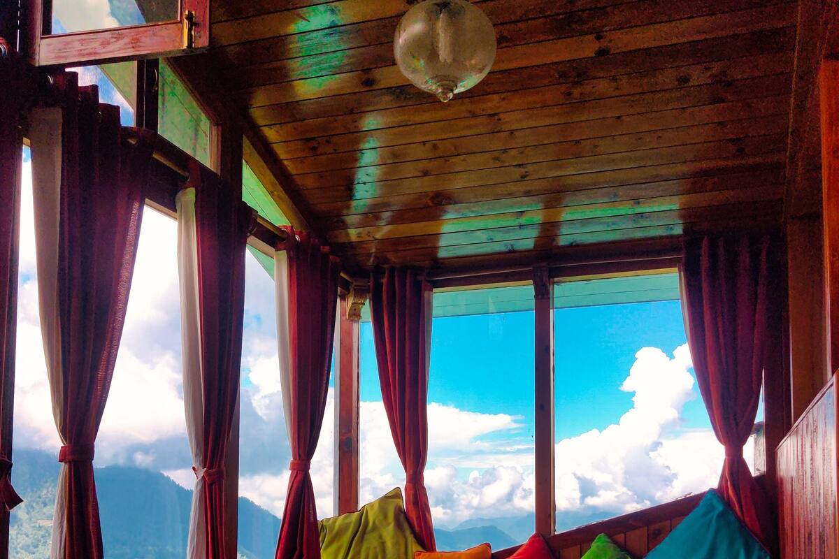 Une cabane en bois avec un banc et des coussins, de grandes fenêtres avec des rideaux et un luminaire suspendu.