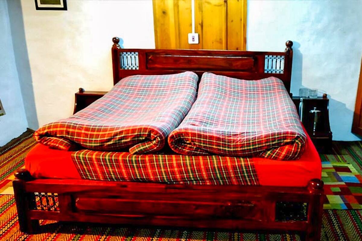 Un lit en bois avec une couvre-lit à carreaux et des taies d’oreiller assorties, une table de chevet avec une lampe, et un tapis à motifs sur le sol.