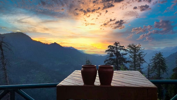 Une table en bois, ornée de deux pots en argile, sur un balcon donnant sur un paysage montagneux au coucher du soleil.