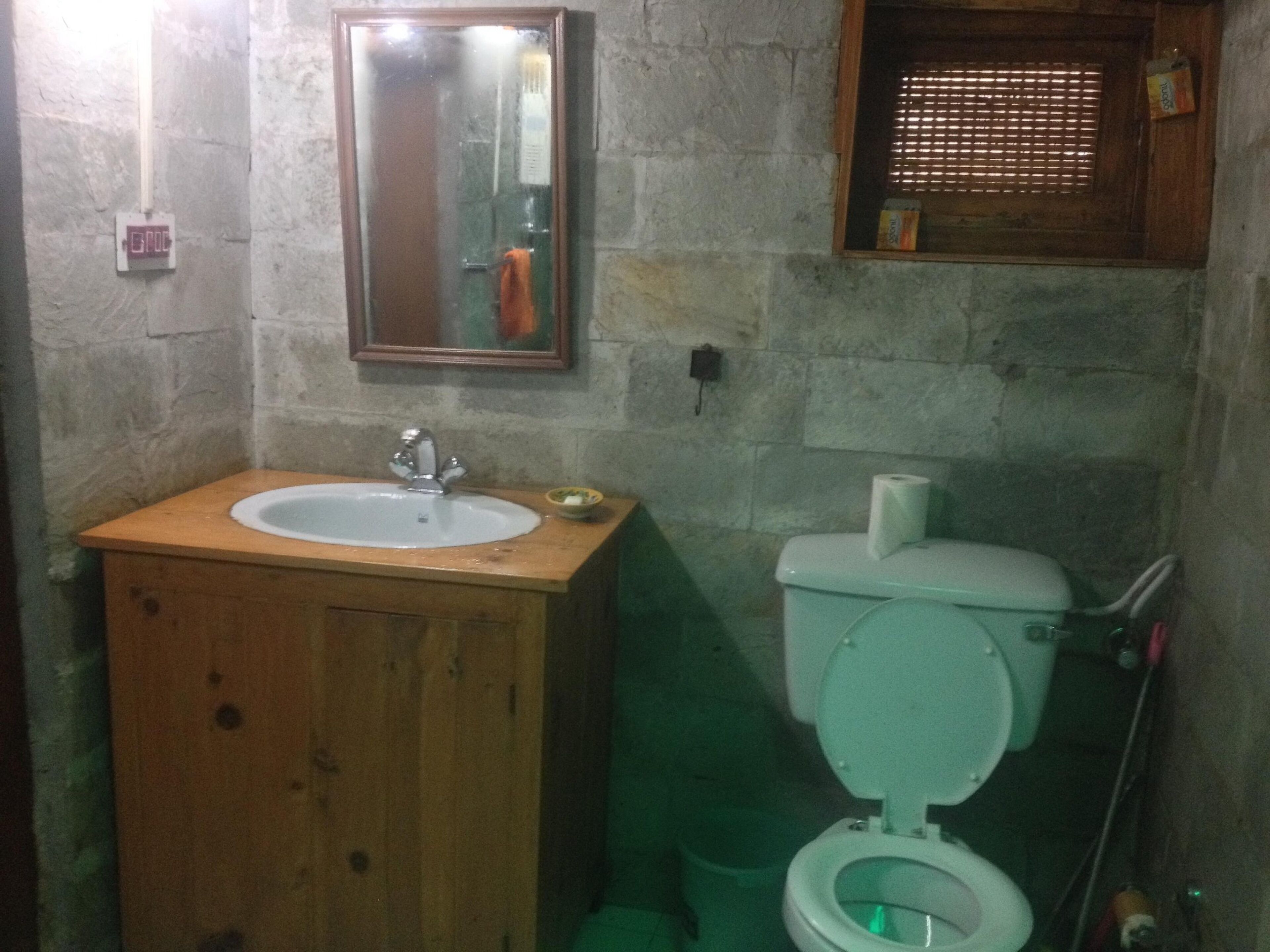 A bathroom with a wooden vanity, a sink, a mirror, a toilet, and a window with blinds.