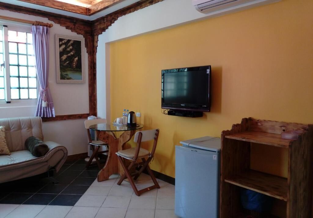 A room with a sofa, a table with chairs, a television mounted on the wall, a small refrigerator, and a wooden shelf.