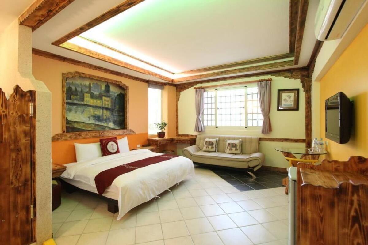 A bedroom with a bed, a sofa, a window with curtains, a painting on the wall, and a TV mounted on the wall.