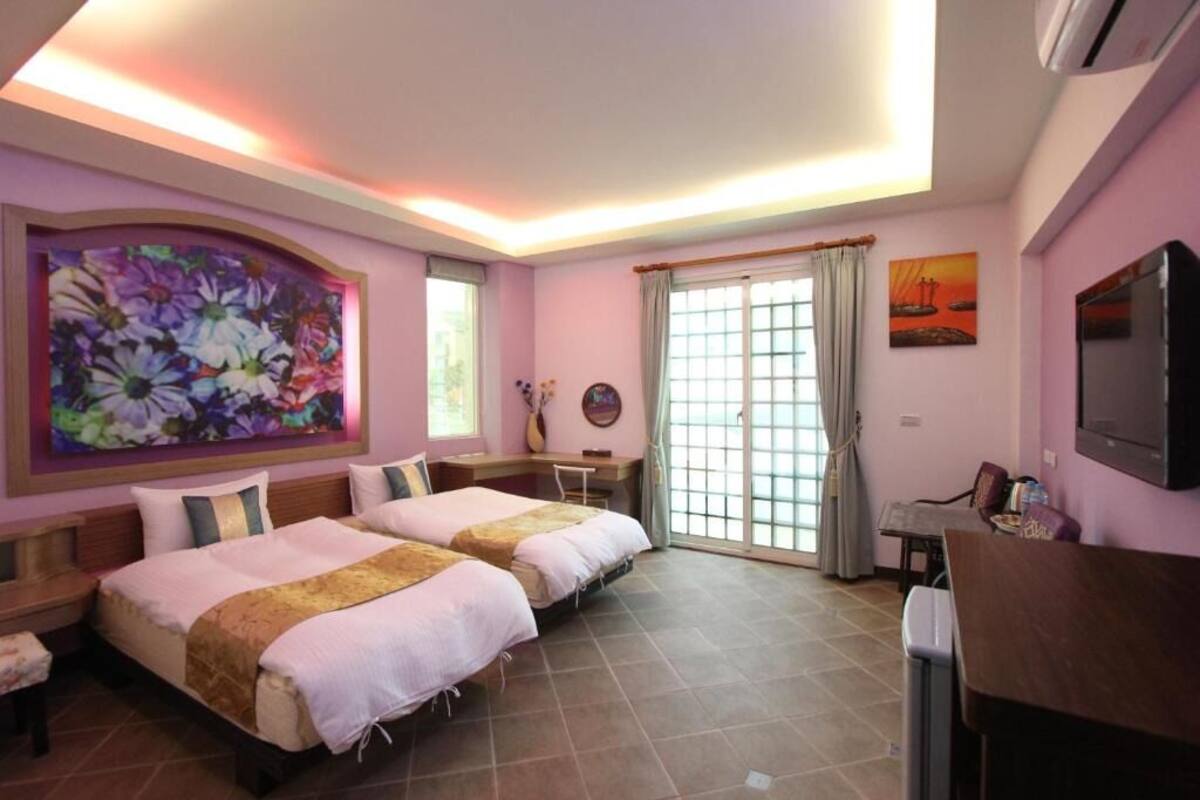 A hotel room with two beds, a desk, a TV, and a large floral painting on the wall.