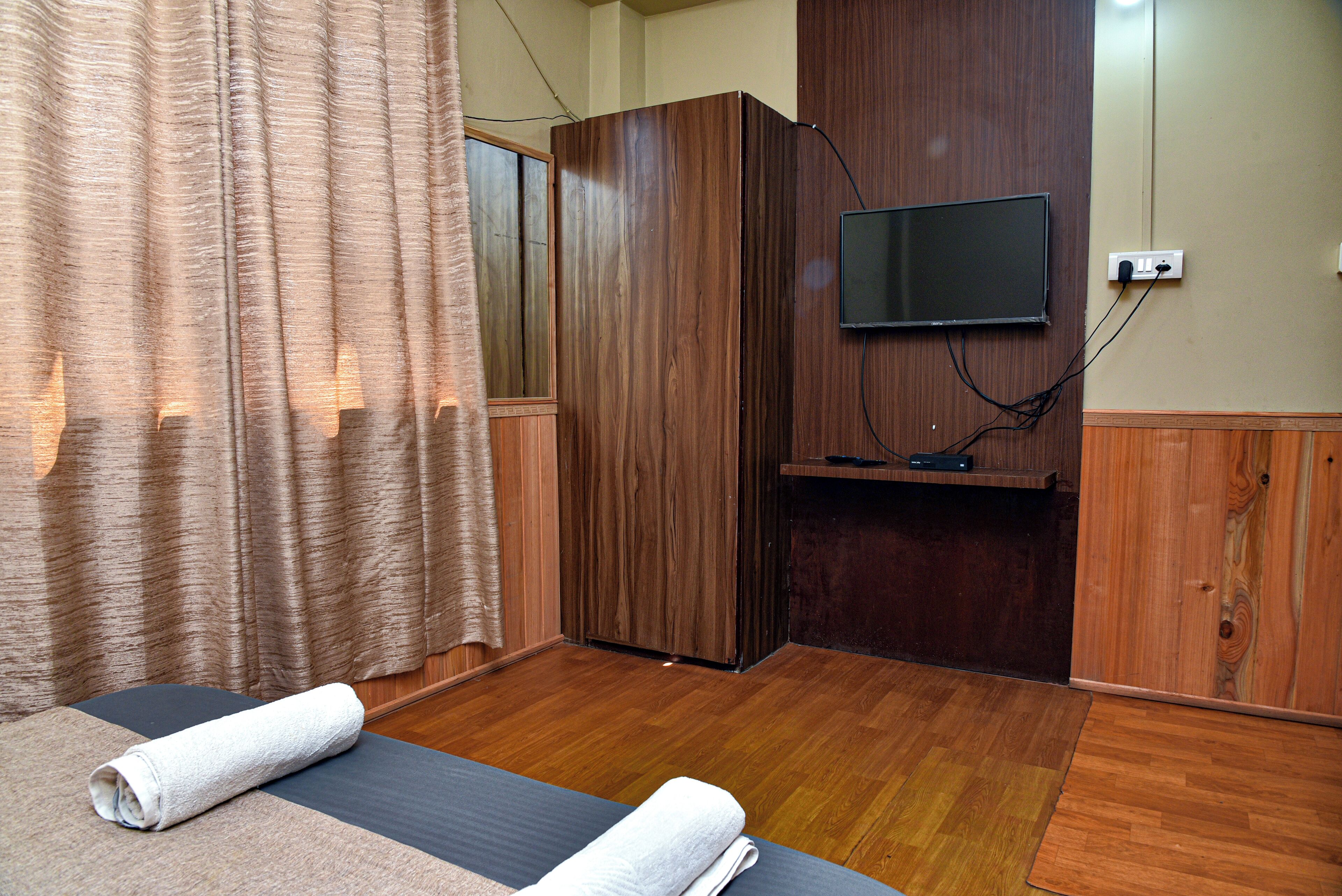 Habitación de hotel con un armario de madera, un televisor de pantalla plana montado en la pared, una cama con una toalla enrollada y suelo de madera.