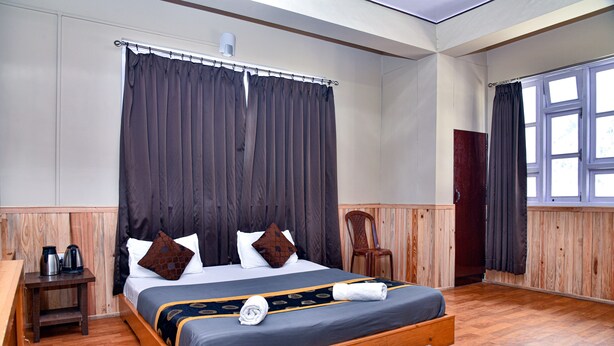 Una cama bien tendida con un cabecero oscuro, una mesita de noche de madera con un hervidor, y una ventana con cortinas.