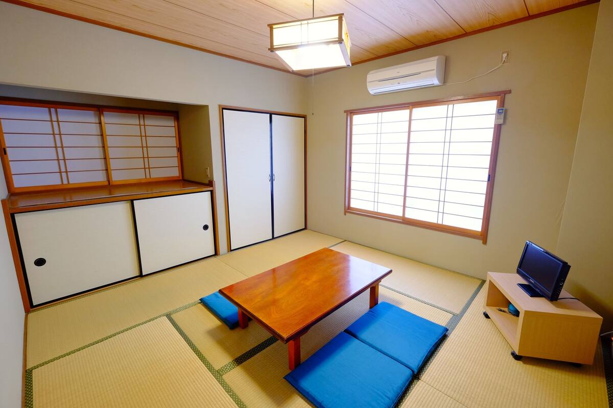 A traditional Japanese room with tatami flooring, a low wooden table, blue cushions, sliding shoji doors, and a television on a wooden stand.