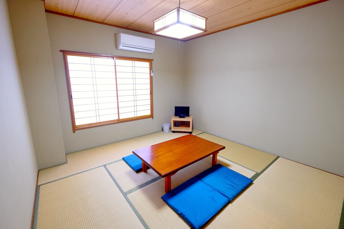 A traditional Japanese room with tatami flooring, a low wooden table, blue cushions, a television, and a ceiling light fixture.