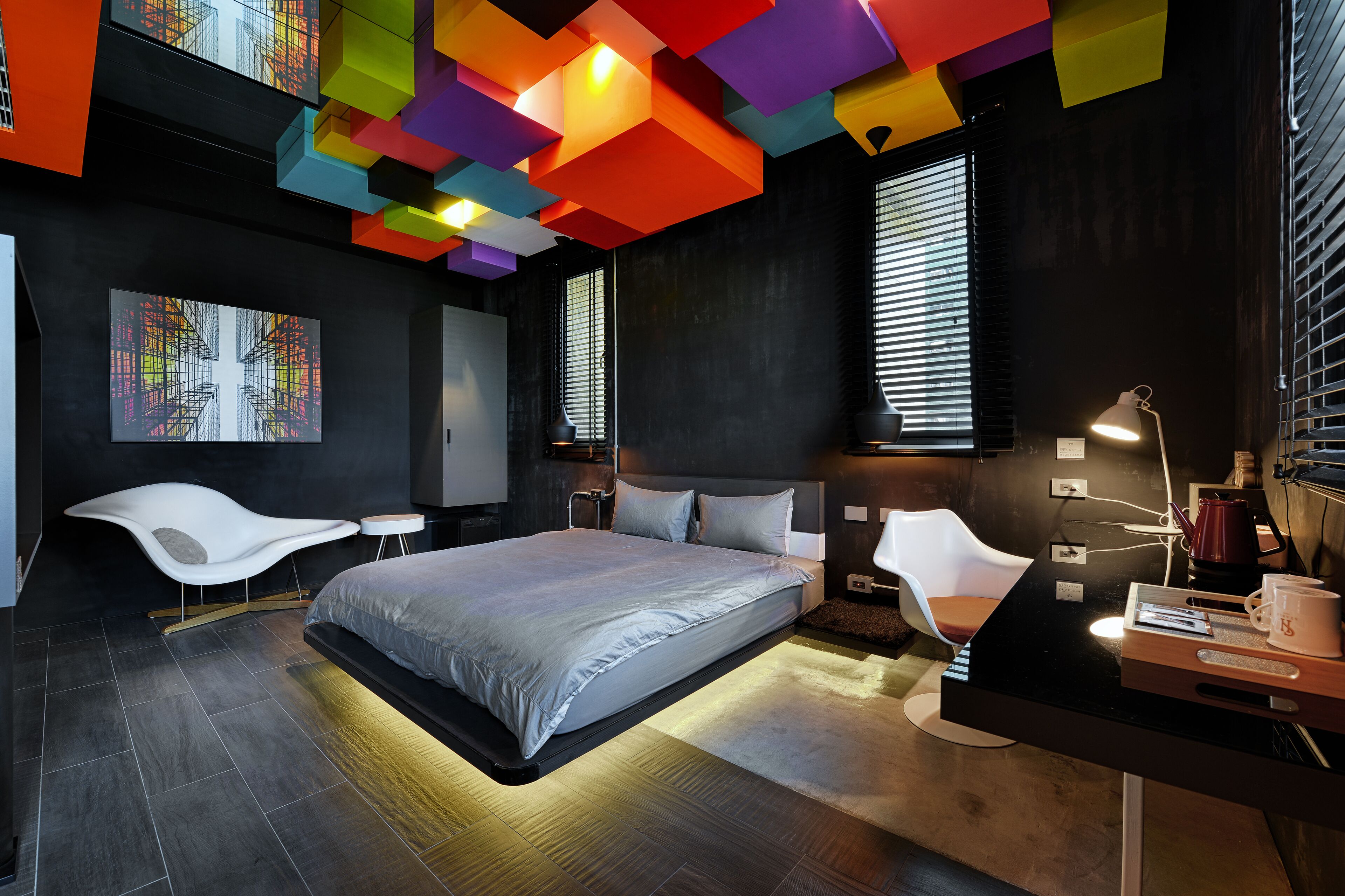 A modern bedroom with a bed, a desk, and a chair. There are windows with blinds, a painting on the wall, and a colorful ceiling.