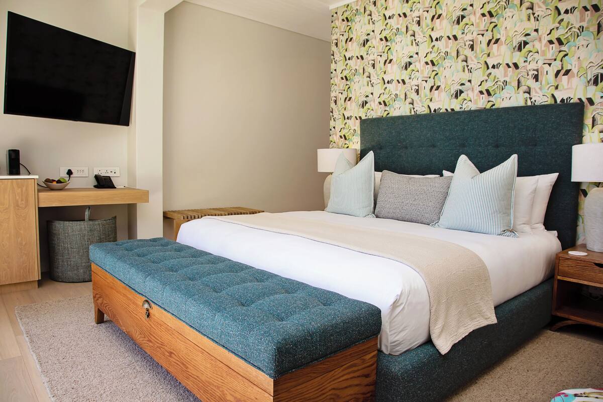 A modern hotel room with a large bed, a flat-screen TV mounted on the wall, a bedside table with a lamp, and a small desk with a bowl of fruit.