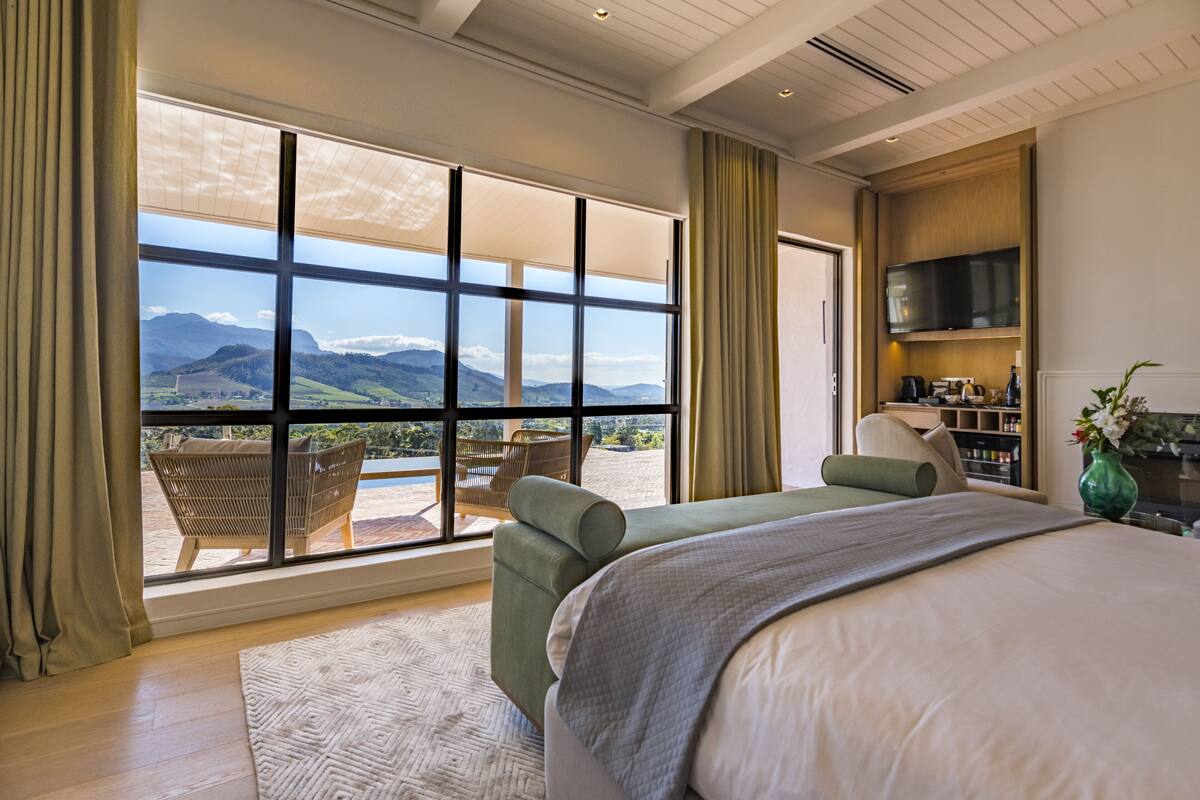 A bedroom with a large window, a bed, a seating area, and a TV.