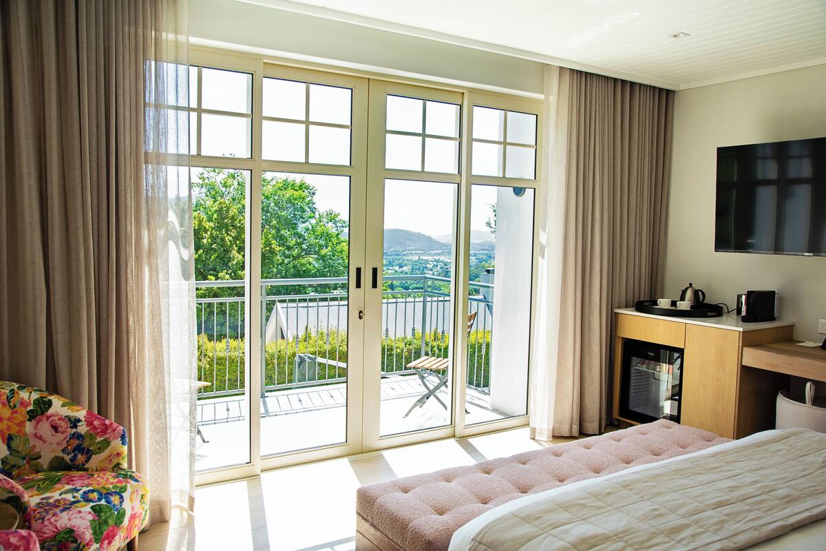 A hotel room with a bed, a desk with a flat-screen TV, a balcony with a view of greenery and hills, and a sofa.