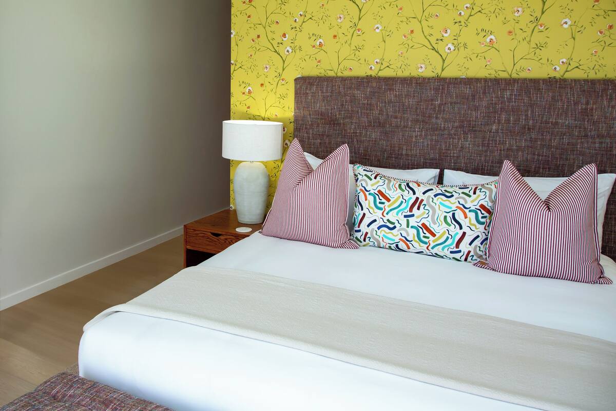 A neatly made bed with a white comforter and multiple pillows, a bedside lamp, and a floral wallpaper pattern on the wall.