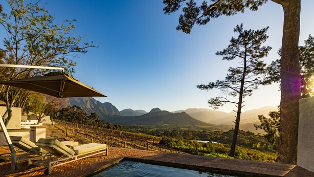 Área de piscina com espreguiçadeiras e um pergolado, cercada por um vinhedo e montanhas ao fundo.