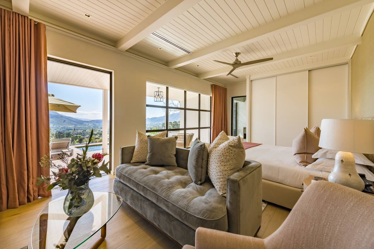 A modern hotel room with a large window offering a mountain view, a sofa, a bed, and a ceiling fan.