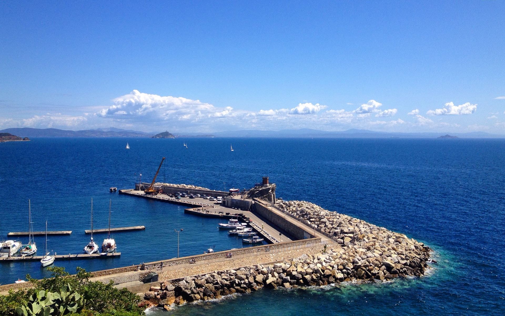 A coastal marina with boats docked, a rocky breakwater, and a clear blue sky.