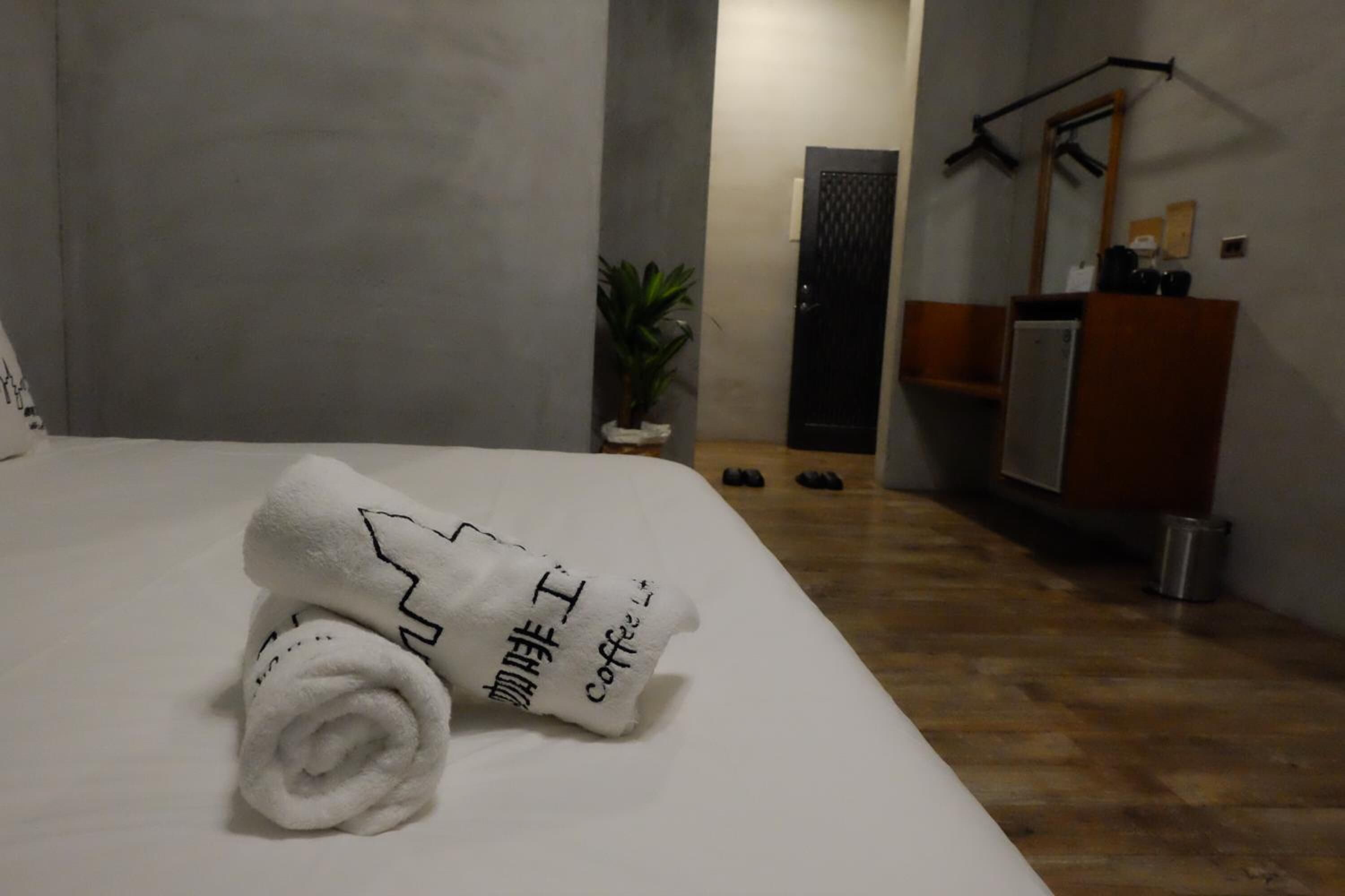 Een netjes opgemaakt bed met een opgerolde witte handdoek met tekst, een potplant, een houten vloer en een houten dressoir met spiegel.