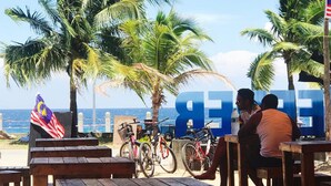 Um café à beira-mar com mesas e bancos de madeira, bicicletas e a bandeira da Malásia.