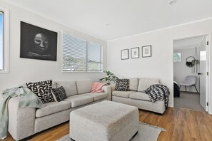 A modern living room with a grey sofa, a matching ottoman, and a large window with blinds. There are framed pictures on the wall and a plant in the corner.