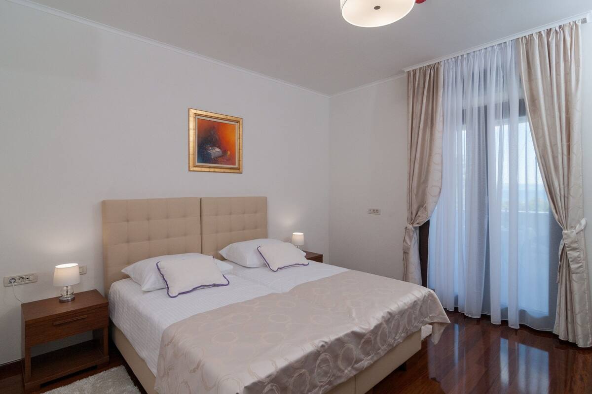 A bedroom with a large bed, two bedside tables, a night lamp, a framed picture on the wall, and sheer curtains on the window.