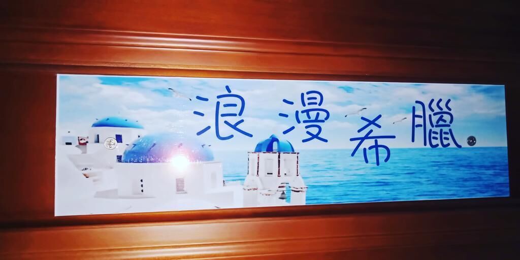 Schild mit Blick auf eine Küstenlandschaft, mit weißen Gebäuden und blauen Kuppeln sowie dem Meer im Hintergrund.