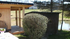 Un belvédère en bois avec un banc et une table, un petit abri de jardin et un étang, le tout intégré dans un cadre verdoyant.