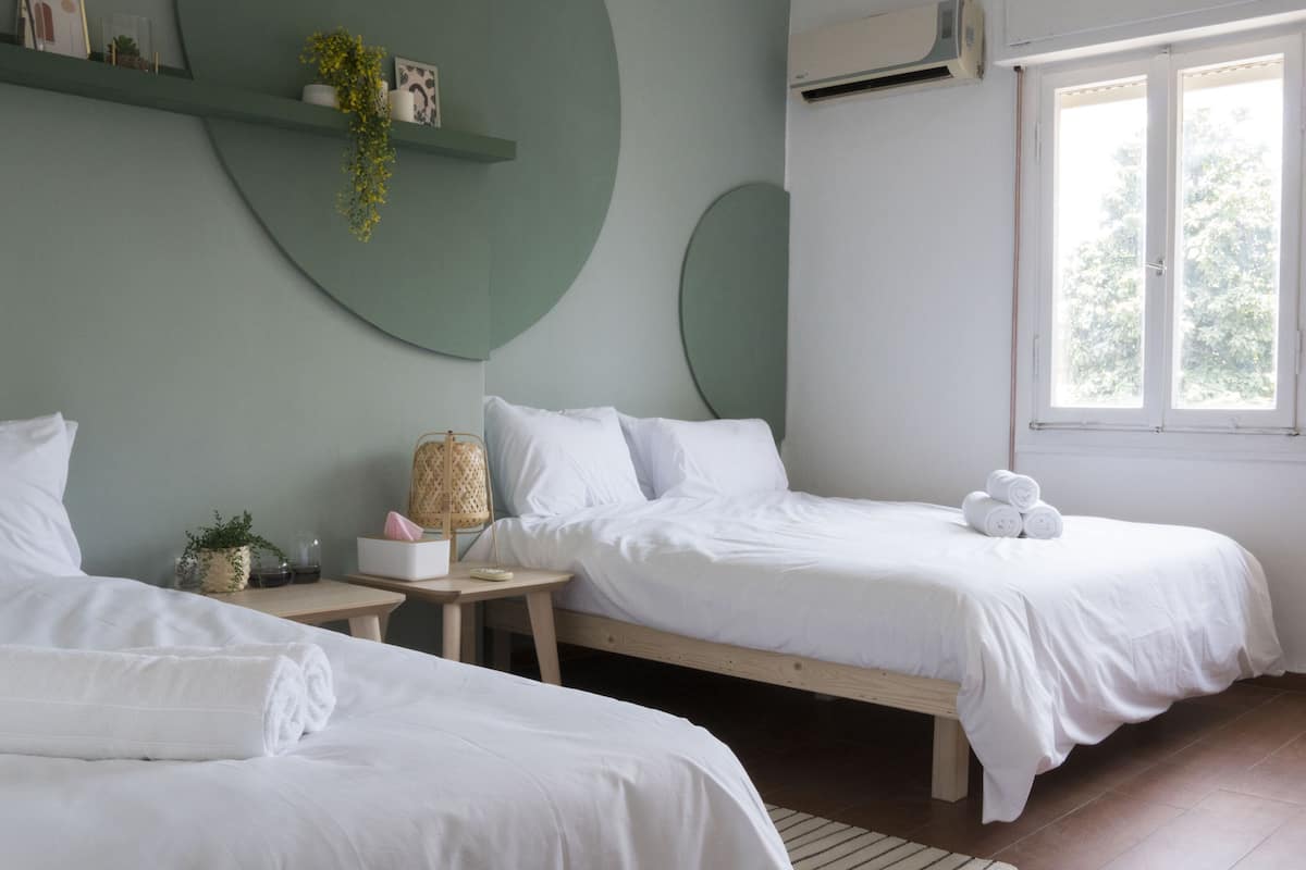 A bedroom with two beds, a bedside table, a hanging plant, and a window with a view of trees.