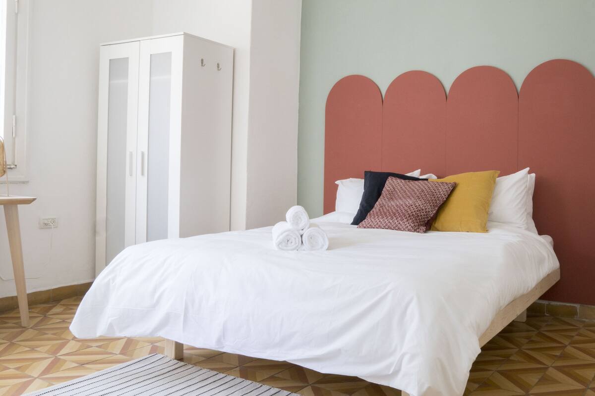 A neatly made bed with white linens, a headboard with a scalloped design, and a bedside table with a framed picture and two candles.