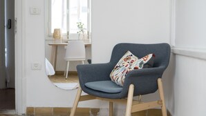 Un sillón gris con un cojín estampado en una habitación con paredes blancas, suelo de madera y una silla blanca al fondo.