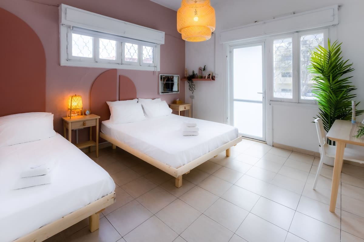 A room with two single beds, a wooden table, a chair, a hanging light fixture, and a window with a view of buildings.