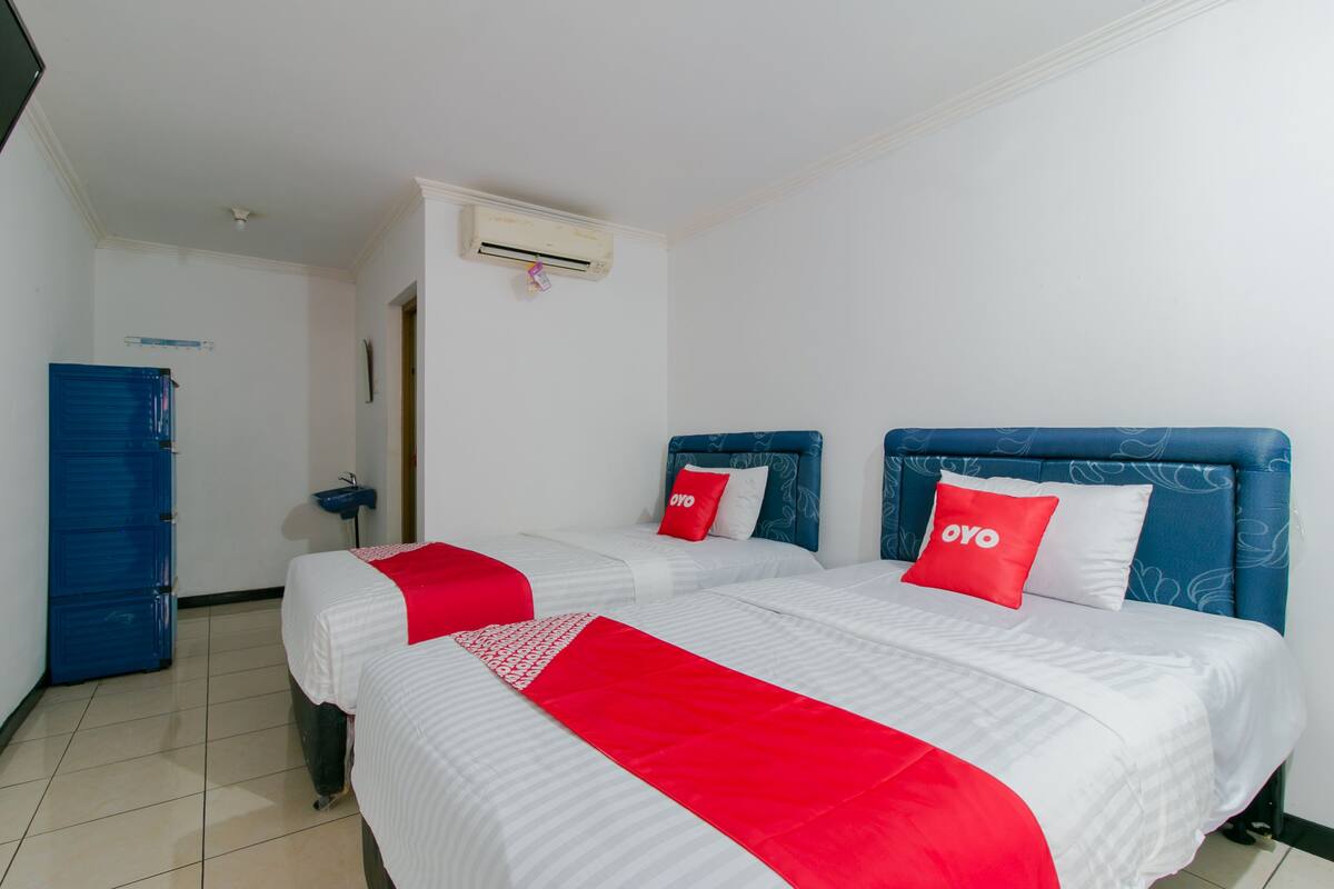 Habitación de hotel con dos camas, cada una con cabecero azul y almohadas rojas, ropa de cama blanca y una manta roja. Se observa un armario azul y una mesita de noche con una lámpara.