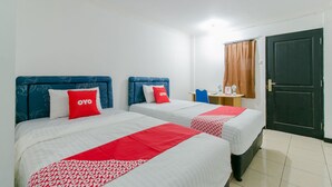 Habitación de hotel con dos camas, cada una con cabecero azul, ropa de cama blanca y detalles rojos. Hay una pequeña mesa con una silla, una ventana con cortinas y una puerta.