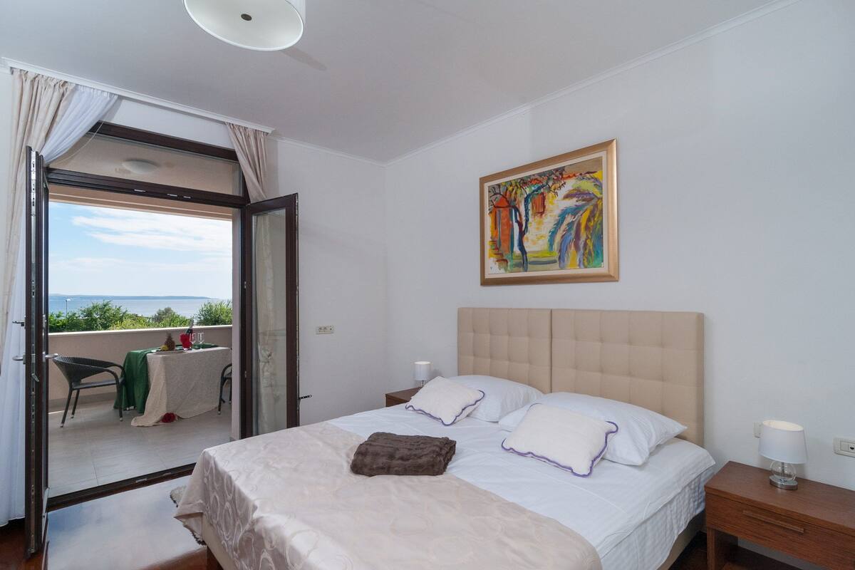 Un dormitorio con cama, mesita de noche, lámpara, un cuadro y vistas a través de un ventanal.