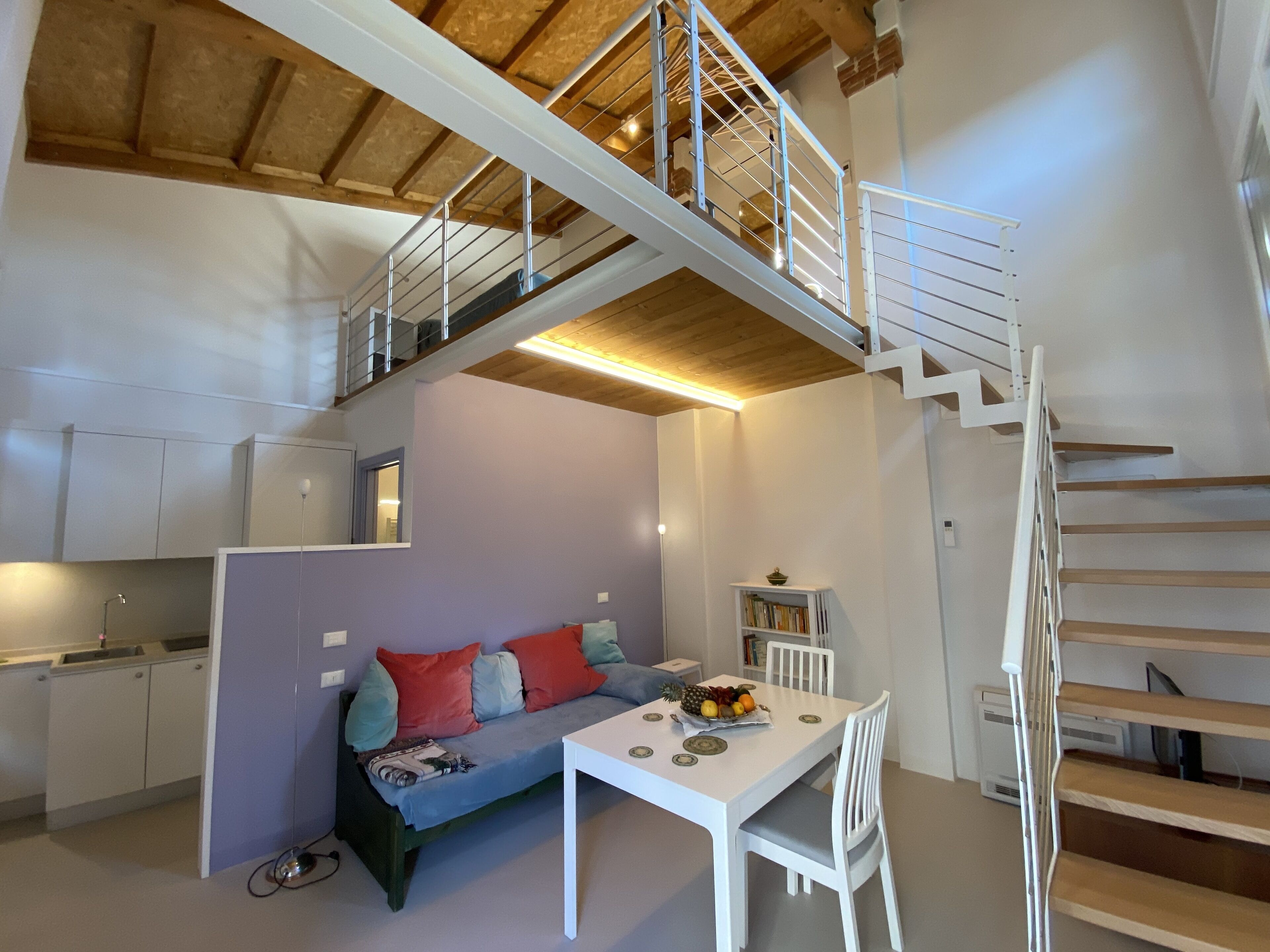 Eine moderne Küche mit weißen Schränken, einem Esstisch mit Stühlen, einer Couch mit bunten Kissen und einer Treppe, die in einen oberen Bereich führt.