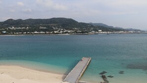 A coastal view with a pier extending into the water, a sandy beach, and a town in the background with buildings and hills. - Leceb Sesoko Villa (Motobu)