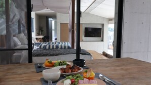 A dining table with breakfast items, a bed with a blue and white striped bedspread, and a view of a bedroom with a large window. - Leceb Sesoko Villa (Motobu)