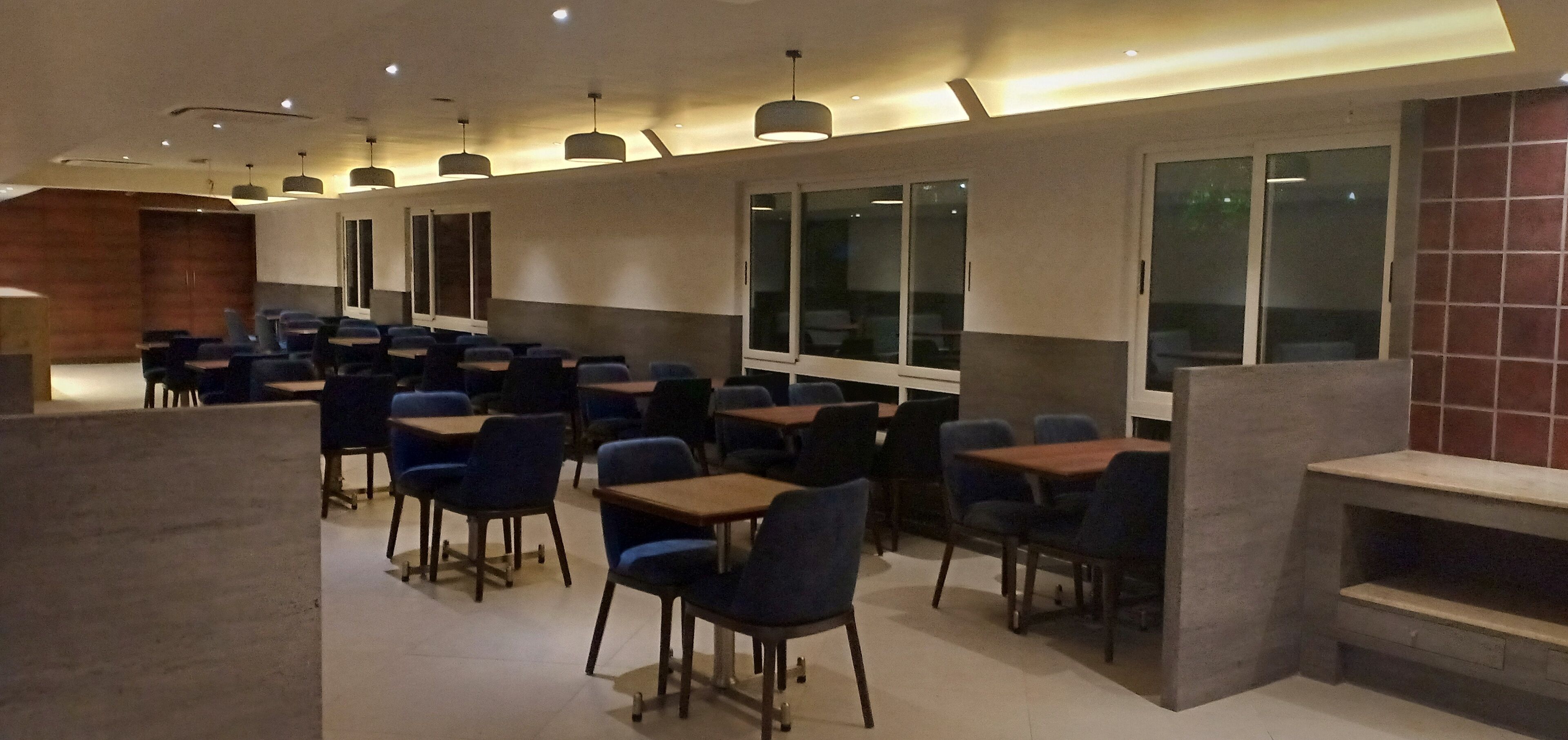Un comedor moderno con mesas y sillas, ventanas amplias e iluminación colgante.