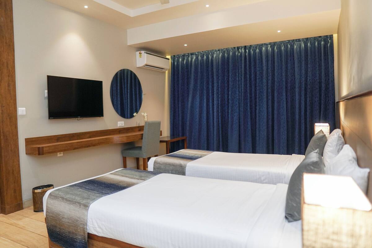 A hotel room with two beds, a TV, a desk, and a large window with curtains.