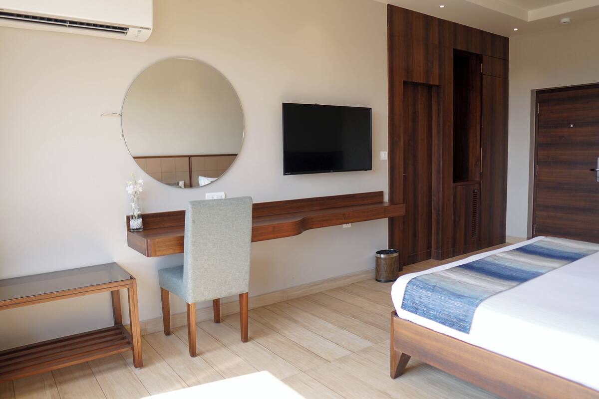 A modern hotel room with a wooden desk, a chair, a bed with a blue and white bedspread, a round mirror, and a flat-screen TV mounted on the wall.