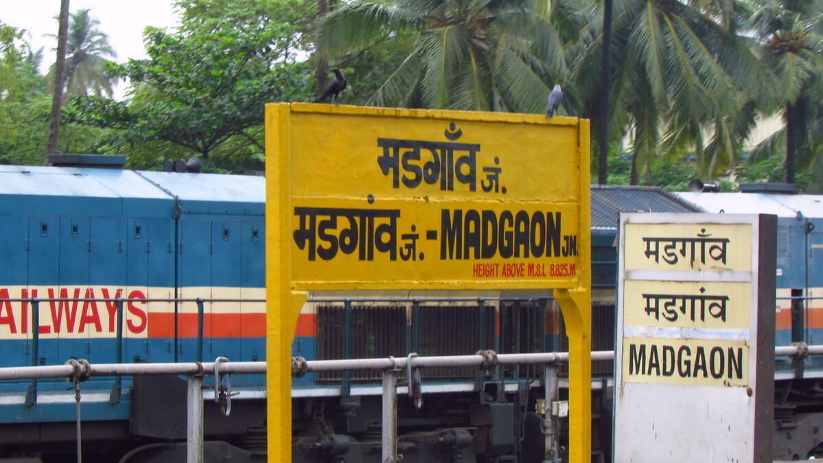 Un andén de estación de tren con un letrero que indica 'Madgaon' y un tren azul con la palabra 'Railways' escrita en él.