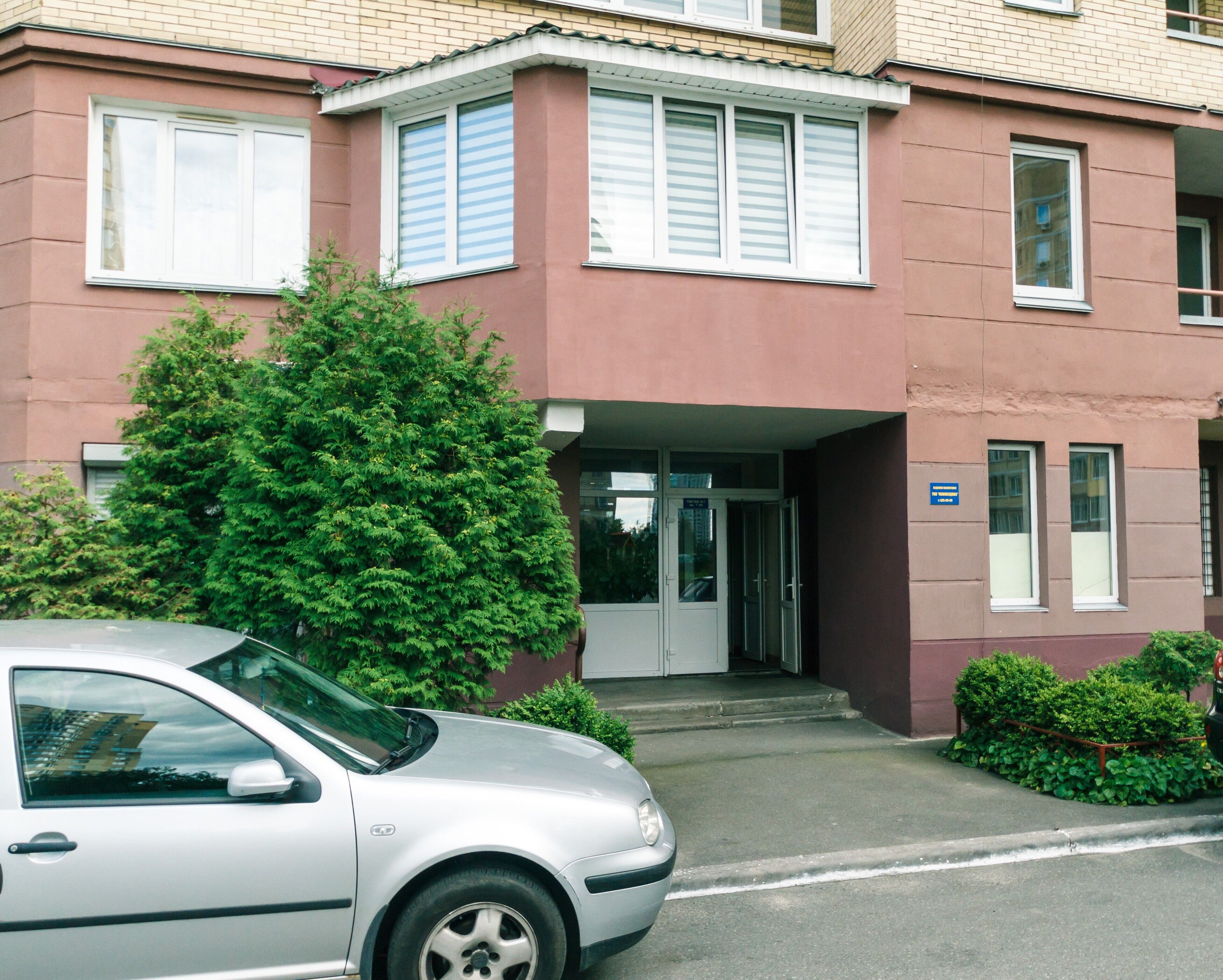 Un bâtiment à deux étages avec une façade rouge, une voiture garée devant et un arbuste vert au premier plan.